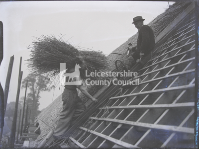 Thatcher delivering large thatch bundle to other man on roof frame	