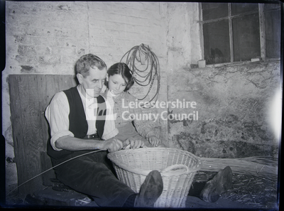 Older man seated in shop with little girl to right, making a basket			