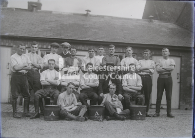 Group portrait of seated stablehands with building behind	