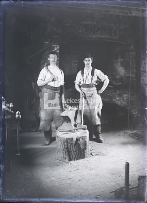 2 smiths standing over anvil holding horseshoe with tongs	