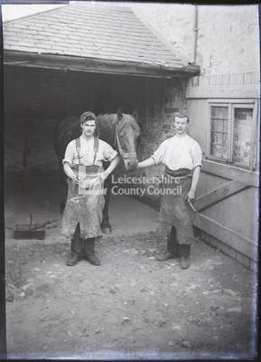 2 men standing on either side of horse in stable yard
