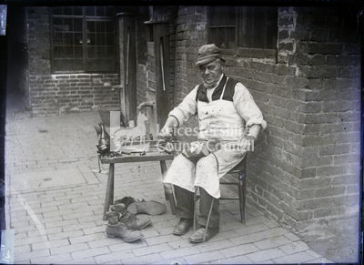 Cobbler with white apron seated at small table with supplies	